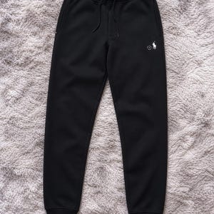 May include: Black sweatpants with a drawstring waist and a small white logo on the left leg. The pants have a relaxed fit and cuffed ankles. The logo features a polo player on a horse.