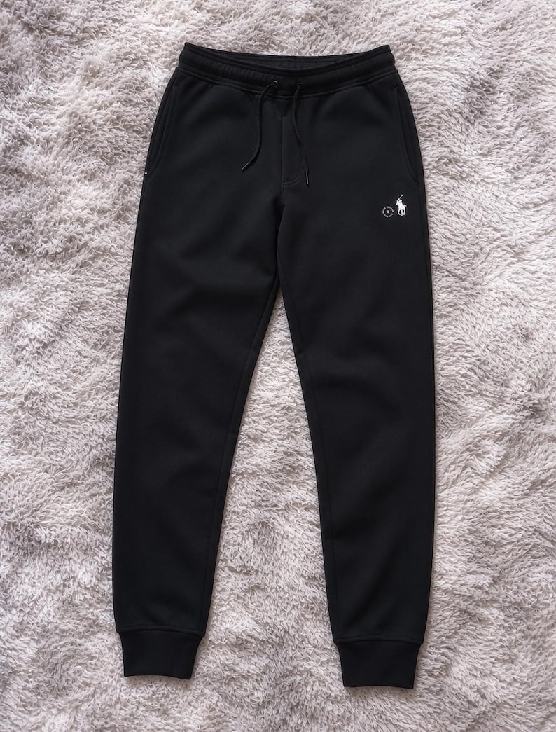 May include: Black sweatpants with a drawstring waist and a small white logo on the left leg. The pants have a relaxed fit and cuffed ankles. The logo features a polo player on a horse.