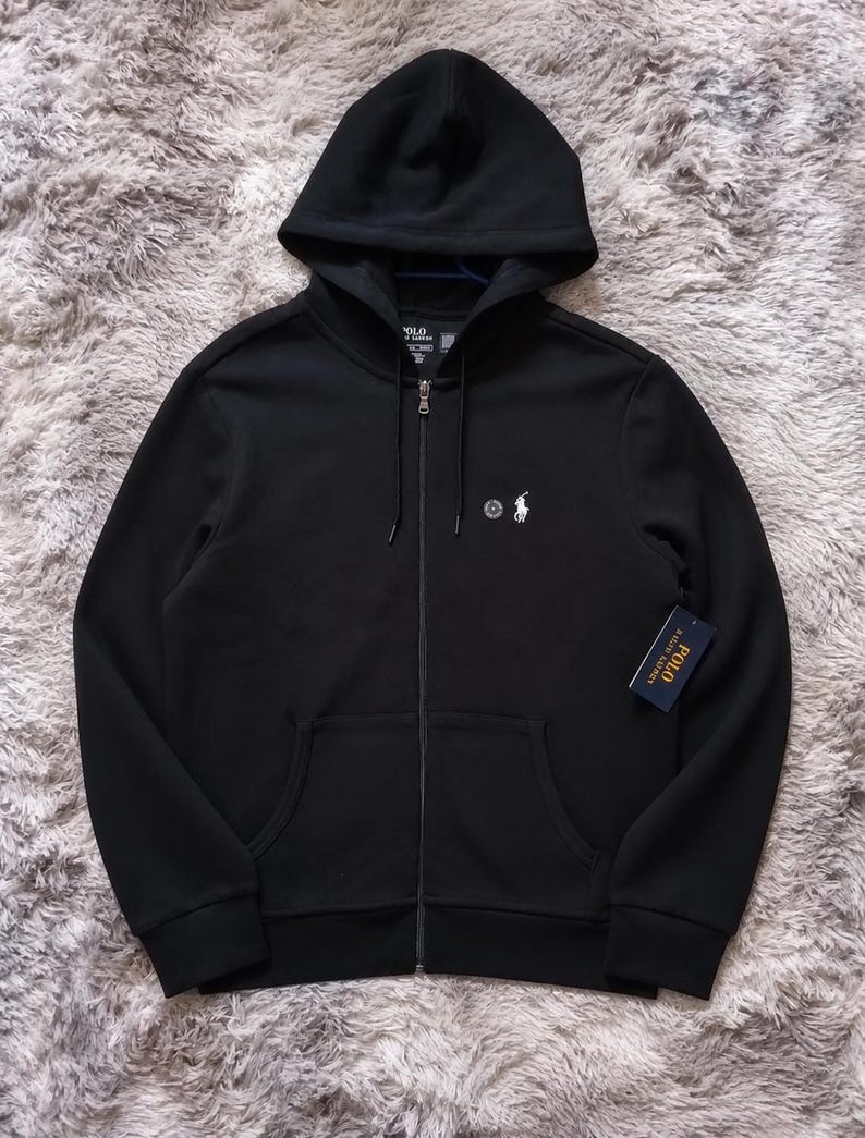 May include: Black zip-up hoodie with a hood and front pockets. Features the Polo Ralph Lauren logo on the chest and a tag on the lower right. The hoodie has a full zipper and a drawstring hood.