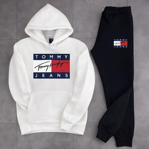 Peut inclure: Un ensemble de survêtement blanc et noir. Le sweat à capuche blanc présente une poche avant et un logo rectangulaire avec le texte "TOMMY JEANS" et la signature Tommy Hilfiger en rouge, blanc et bleu. Le pantalon de survêtement noir a un logo assorti.