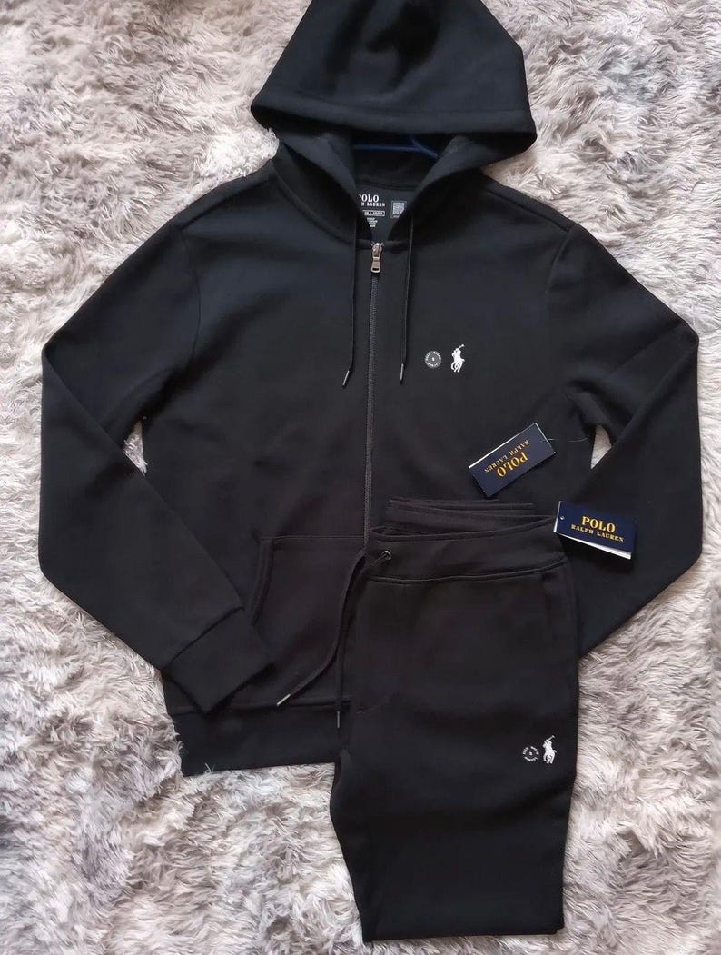 May include: Black hooded sweatshirt and matching sweatpants set. The sweatshirt has a full zipper, front pockets, and a small logo on the chest. The sweatpants have a drawstring waist and a matching logo. The set is displayed on a textured surface.