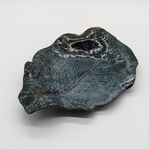 May include: A ceramic dish shaped like a large leaf with an irregular, wavy edge. The surface features a dark blue and black glaze with a glossy finish, highlighting the intricate, raised vein patterns that extend across the entire structure.