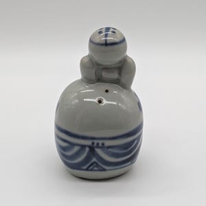 May include: A ceramic salt shaker in the shape of a seated figure. The shaker is primarily gray with blue decorative bands and accents. The figure's head has a blue cross design. The shaker has three holes for dispensing salt.
