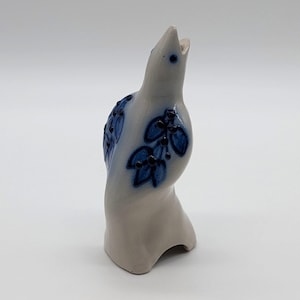 May include: A ceramic bird figurine in a light gray color with blue floral designs. The bird has an open beak and is standing upright. The blue design includes leaves and berries. The figurine is a decorative item.