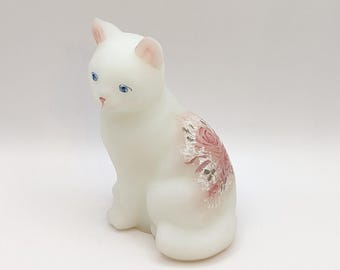 Puede incluir: Una figura de gato de vidrio lechoso blanco en posición sentada. El gato presenta ojos azules pintados, orejas internas rosadas y una nariz rosada. Un patrón floral decorativo con rosas rosadas y detalles plateados está pintado en el costado.