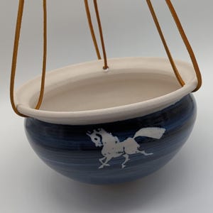 May include: A hanging ceramic planter with a navy blue exterior and a white interior. The planter features a white horse silhouette and is suspended by brown leather straps. The design is simple and elegant, suitable for indoor or outdoor use.