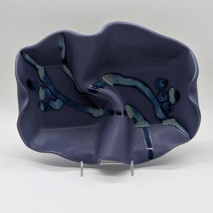 May include: A matte purple ceramic tray with a wavy, organic rim and a central fold. The surface features a decorative pattern of dark blue and light blue painted branches with small circular accents, creating a textured, artistic appearance.