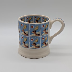 May include: A ceramic mug with a cream-colored base and handle, featuring a repeating pattern of postage stamp-style illustrations. Each stamp depicts a brown deer with large antlers against a light blue background with a small red circle.