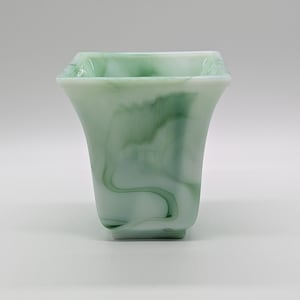 May include: A square-shaped glass vase featuring a marbled pattern of translucent white and soft green swirls. The vessel has a wide, flared rim that tapers down to a smaller, flat base, creating a smooth, polished finish throughout the object.