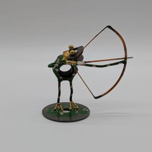 May include: A metal sculpture of a frog with a camouflage pattern, featuring gold-toned eyes and limbs. The frog stands on a circular base and holds a copper-colored bow and arrow, with a large nut forming the central body of the figure.