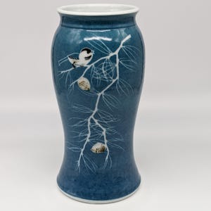 May include: A blue ceramic vase with a white interior rim, featuring a hand-painted design of a small bird perched on a pine branch with two pinecones. The branch and needles are rendered in white, contrasting against the mottled blue background.