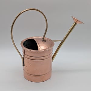 May include: A copper watering can with a brass handle and spout. The can has a cylindrical body with horizontal ridges and a curved handle. The spout is angled and has a small, flat head. The can is designed for watering plants.