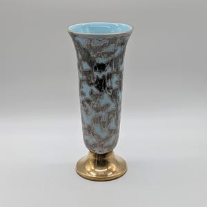 May include: A tall, slender vase featuring a light blue interior and a marbled exterior pattern of dark brown and light blue swirls. The vase is supported by a polished, reflective gold-toned circular metal base that flares outward at the bottom.