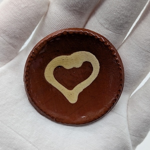 May include: A small, round, brown ceramic dish with a cream-colored heart design in the center. The rim of the dish has a textured, scalloped edge. The dish is handmade and has a rustic appearance.