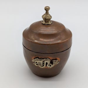 May include: A small, rounded copper container with a matching lid featuring a metallic finial knob. The body of the vessel is adorned with a silver-toned metal emblem depicting a dragon figure, centered on the front surface of the copper jar.