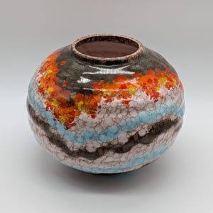 May include: A round ceramic vase with a wide opening. The vase features a textured surface with a color palette of brown, orange, light blue, and white. The top rim is dark brown.