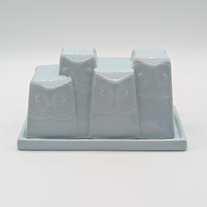 May include: A light blue ceramic butter dish featuring a rectangular base and a lid molded with the relief design of four stylized owls standing side by side. The glossy finish highlights the embossed circular eyes and feathered textures of the owls.