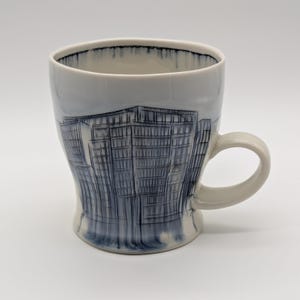 May include: A white ceramic mug with a curved handle. The mug features a blue and gray architectural illustration of a building. The rim of the mug has a dark blue trim.