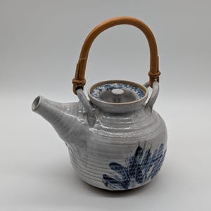 May include: A ceramic teapot with a light gray glazed finish and a curved bamboo handle. The body features a textured, ribbed surface with a blue painted leaf-like pattern near the base. It includes a matching lid with a small central knob.