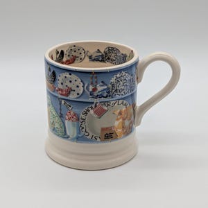 May include: A ceramic mug with a cream-colored base and a blue exterior featuring a repeating pattern of various plates, teapots, and text including "Village Hall" and "Shoulder of Lamb". The interior rim also displays a pattern of small plates.
