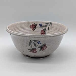 May include: A speckled off-white ceramic colander bowl featuring a hand-painted floral design with muted red blossoms, green leaves, and small blue accents. The bowl has a rounded shape with visible drainage holes near the base and a rimmed edge.
