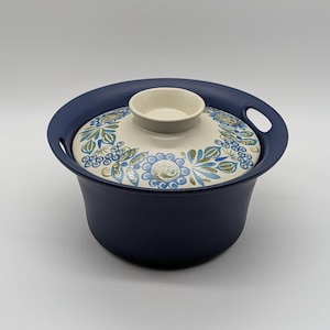 May include: A deep navy blue ceramic pot with a matching lid featuring a cream-colored top surface decorated with a circular pattern of blue and olive green floral motifs. The pot includes two integrated side handles and a central knob on the lid.