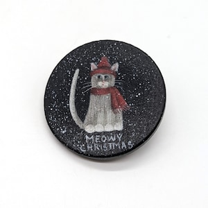 May include: A circular black pin featuring a gray cat wearing a red knit hat and a matching red scarf. The background is black with white speckles resembling falling snow. The text MEOWY CHRISTMAS is printed in white at the bottom of the pin.