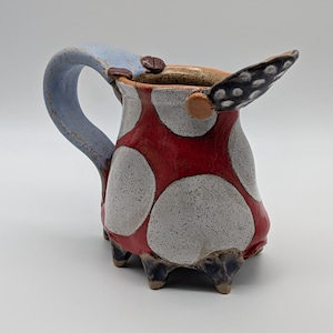 May include: A whimsical ceramic pitcher with a light blue handle and a red body adorned with large white polka dots. The pitcher has a unique wing-like detail and rests on three small feet.