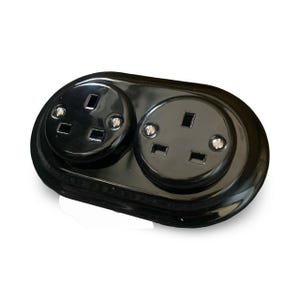Può includere: Una presa elettrica doppia nera con due prese circolari. Ogni presa ha tre aperture rettangolari ed è inserita in una cornice ovale lucida. La presa è progettata per l'uso elettrico.