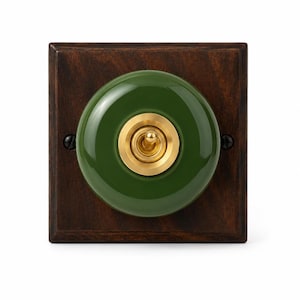 Puede incluir: Una placa de pared cuadrada de madera marrón oscuro con un centro circular de cerámica verde bosque brillante. Un interruptor de palanca central de latón está montado dentro del círculo de cerámica verde, fijado con dos tornillos negros a los lados.