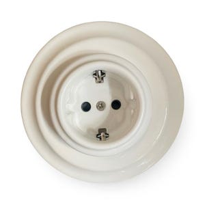 以下が含まれることがあります： 白のセラミック製電気コンセント。円形のデザインで、2つの黒い穴と中央のネジがあり、同心円状のリングで囲まれています。 クリーンでモダンなデザインで、インテリアに最適です。