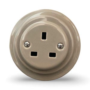 Puede incluir: Una toma de corriente redonda de cerámica gris claro con tres clavijas. La toma tiene un borde ligeramente elevado y dos tornillos plateados.