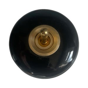 May include: A round, black light switch with a brass toggle and a brass-colored ring around the toggle. The switch has a glossy finish and is set against a white background.