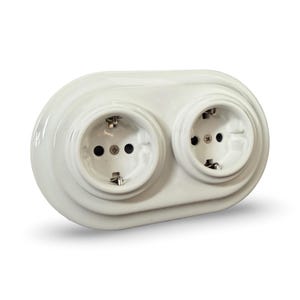 Może przedstawiać: Białe, podwójne gniazdo elektryczne z dwoma okrągłymi gniazdami. Gniazdo wykonane jest z ceramiki i ma błyszczące wykończenie. Gniazda mają po trzy otwory i metalowe styki.