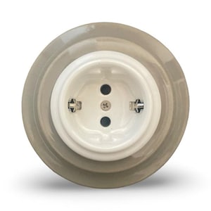 Può includere: Una presa elettrica in ceramica bianca con una cornice in ceramica grigia. La presa ha tre spine ed è progettata per l'uso europeo.