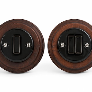 May include: Two circular light switches mounted on dark brown wooden bases. The left switch features a single black toggle, while the right switch has two black toggles. Both units are secured with visible silver-coloured metal screws.