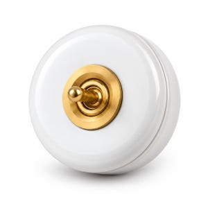 May include: A circular white light switch with a glossy finish, featuring a central brass toggle mechanism. The switch is mounted on a round white base, and the toggle lever is positioned in the centre of a circular brass plate with concentric rings.