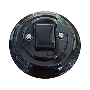 May include: A round, black light switch with a glossy finish. The switch has a rectangular toggle in the center and is secured with two silver screws. The design is a classic, vintage style.