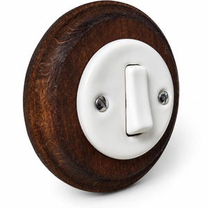Könnte beinhalten: Ein runder Lichtschalter mit einer weißen Keramik-Abdeckplatte auf einem dunkelbraun gebeizten Holzsockel. Die weiße Platte ist mit zwei silberfarbenen Metallschrauben befestigt, die sich auf beiden Seiten des zentralen Kippschalters befinden.