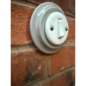 Pode incluir: Um interruptor de luz de estilo vintage com um interruptor branco e um centro branco, embutido numa base de cerâmica cinza e circular. O interruptor é montado numa parede de tijolo vermelho, exibindo um design clássico.