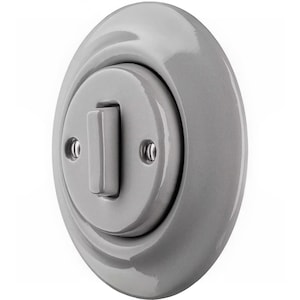 Puede incluir: Un interruptor de luz de cerámica gris con un acabado brillante. El interruptor tiene una forma ovalada redondeada con una palanca rectangular central y dos pequeños orificios para tornillos. El diseño es minimalista y moderno.