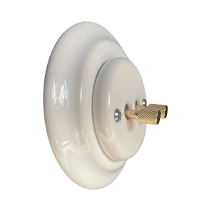 May include: A white ceramic light switch with two brass toggle switches. The switch has a rounded, layered design and is mounted on a white surface. The brass toggles have a textured, knurled finish.