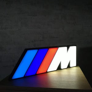 Könnte beinhalten: Beleuchtetes BMW M Logo Schild. Das Schild hat einen schwarzen Rahmen mit blau, rot und weiß beleuchteten Abschnitten. Das M ist weiß und sticht hervor. Das Schild ist auf einer Holzoberfläche angewinkelt. Es ist etwa 30 cm lang.