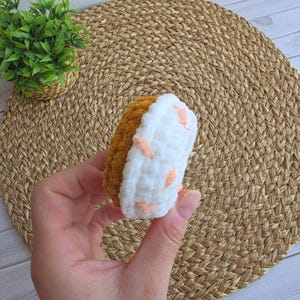 Puede incluir: Un peque&ntilde;o juguete de peluche tejido a ganchillo, redondo, que se asemeja a una rosquilla. La base es de hilo texturizado de color marr&oacute;n dorado, mientras que la parte superior est&aacute; cubierta de hilo de felpa blanco con detalles rosas.