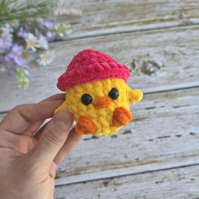May include: A small, handmade crochet plush toy shaped like a yellow chick wearing a bright pink hat. The chick features two round black plastic eyes, a small orange beak, and two orange feet. The texture of the yarn is soft and fuzzy.