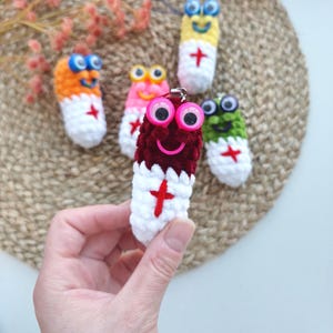 May include: A collection of colorful, crocheted pill-shaped keychains with googly eyes and smiling faces. Each keychain features a different color combination, including orange, pink, yellow, green, and burgundy, with a white base and a red cross design.