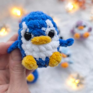 May include: A handmade blue, white, and yellow crochet penguin. The penguin has black eyes, a yellow beak, and yellow feet. Other similar penguins are visible in the background. The penguin is a soft, plush toy.