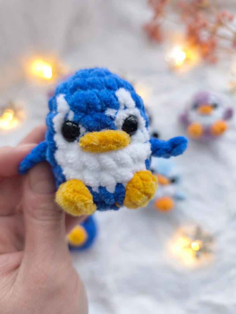 May include: A handmade blue, white, and yellow crochet penguin. The penguin has black eyes, a yellow beak, and yellow feet. Other similar penguins are visible in the background. The penguin is a soft, plush toy.