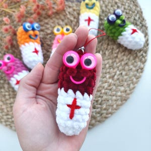 May include: A collection of colorful, handmade crochet ornaments. The main ornament is held in a hand and is a burgundy and white pill shape with a red cross and pink googly eyes. Other ornaments are in various colors.