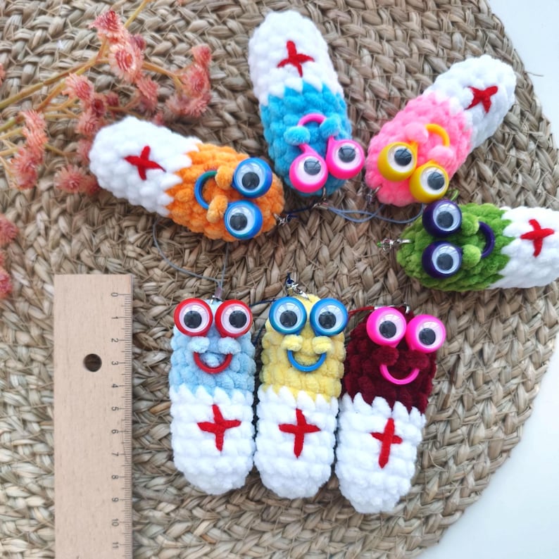 May include: A collection of colorful, handmade crochet pill-shaped keychains. Each keychain features a different color combination, googly eyes, and a red cross design. The keychains are arranged on a woven surface.